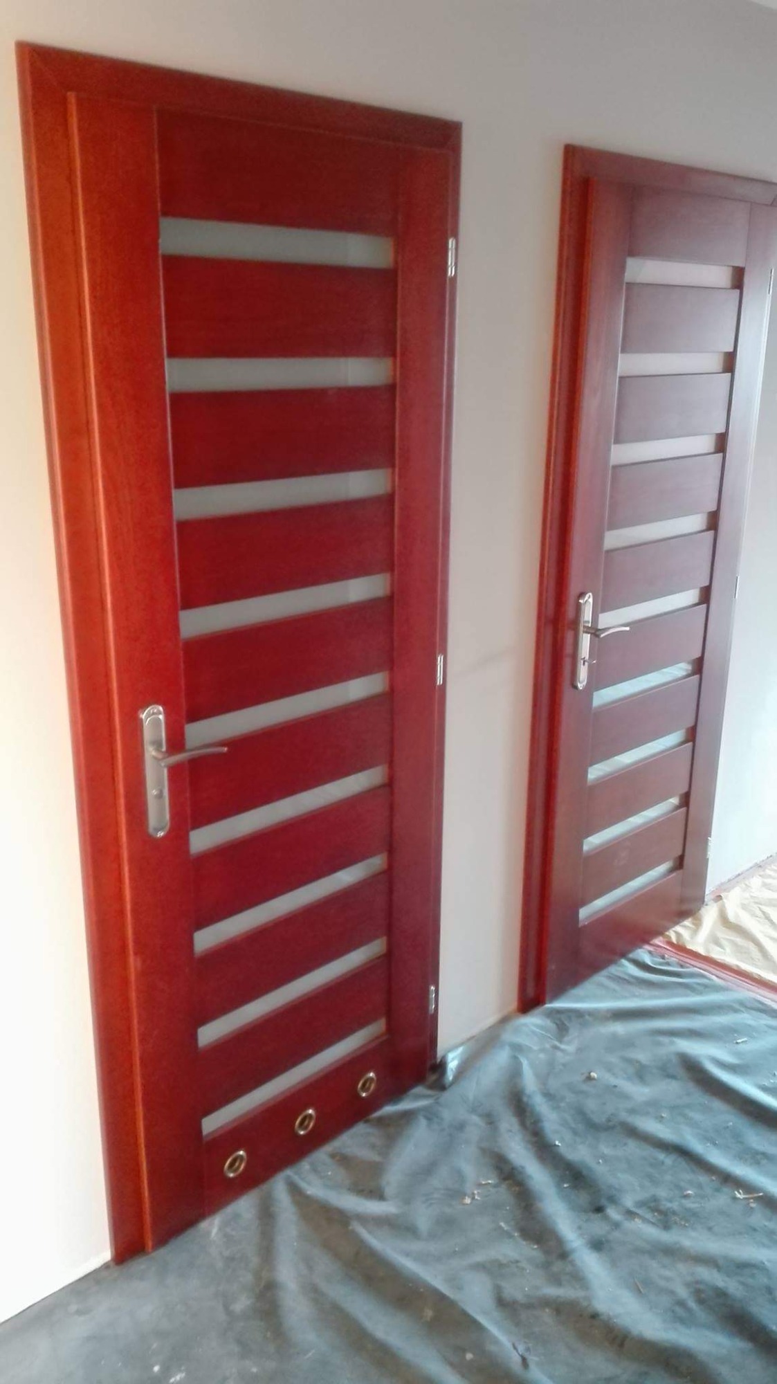 Dwa ciemnoczerwone drzwi wewnętrzne z poziomymi, matowymi przeszkleniami, zamontowane w świeżo pomalowanym pomieszczeniu, zabezpieczonym folią malarską.