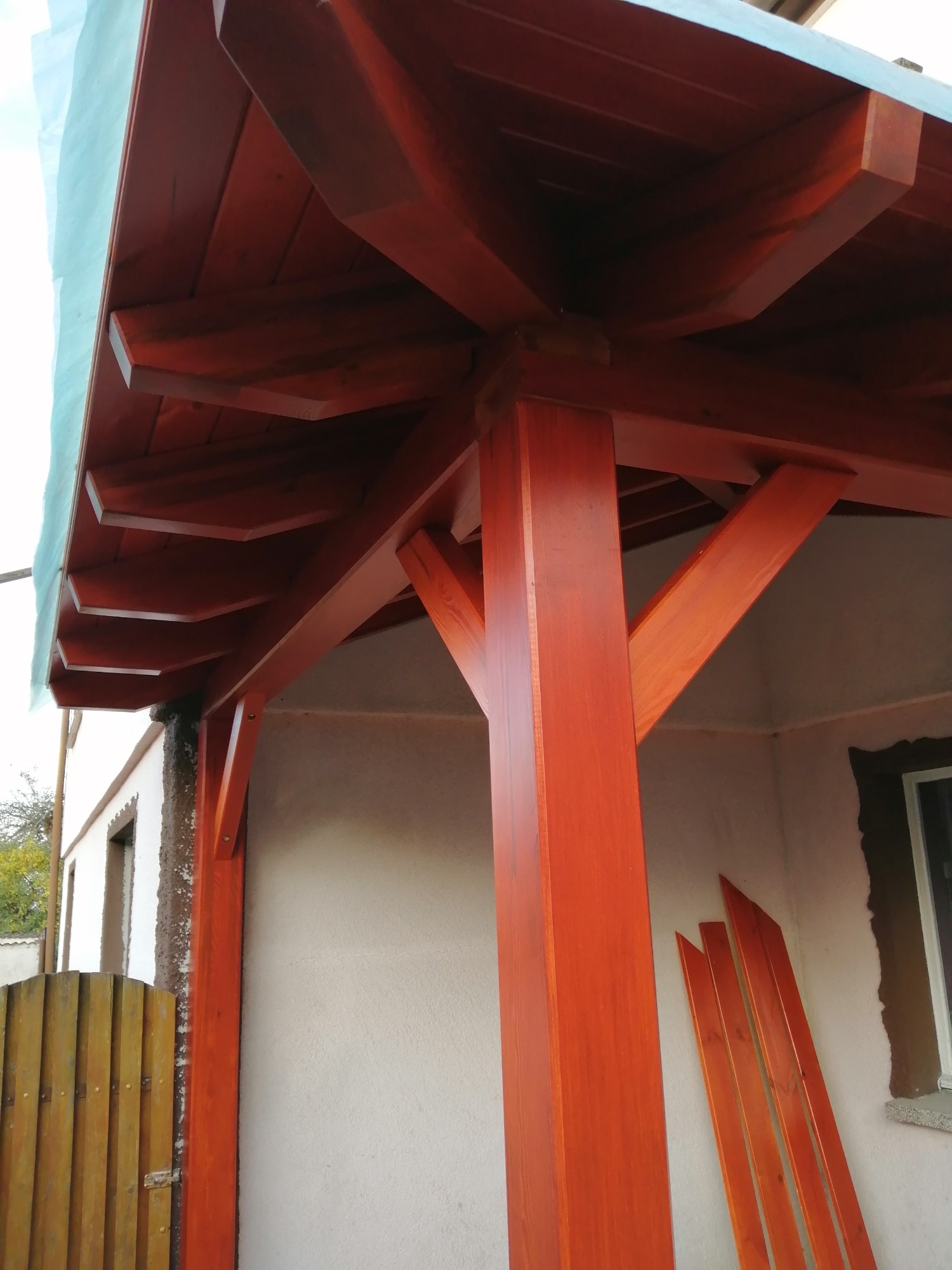 Konstrukcja z belek w kolorze terakoty, prawdopodobnie zadaszenie tarasu, z widocznymi elementami mocującymi i deskami przygotowanymi do montażu przy ścianie budynku.
