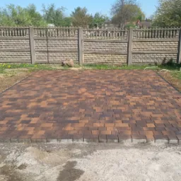 Ukończona nawierzchnia z brązowo-pomarańczowej kostki brukowej ułożonej w jodełkę, obrzeża z szarego betonu, w tle betonowy płot, trawnik z mleczami i drzewa.