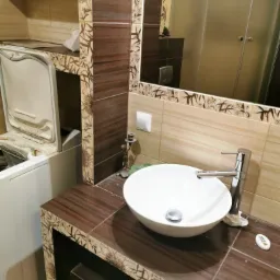 Łazienka z umywalką nablatową, brązowe płytki imitujące drewno na blacie i obudowie lustra, pralka obok umywalki, widoczne niedoróbki wykończeniowe.