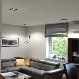 Salon z szarym narożnikiem, drewnianą podłogą i oświetleniem punktowym w suficie, widok na okno z roletą rzymską i zielenią za nim.