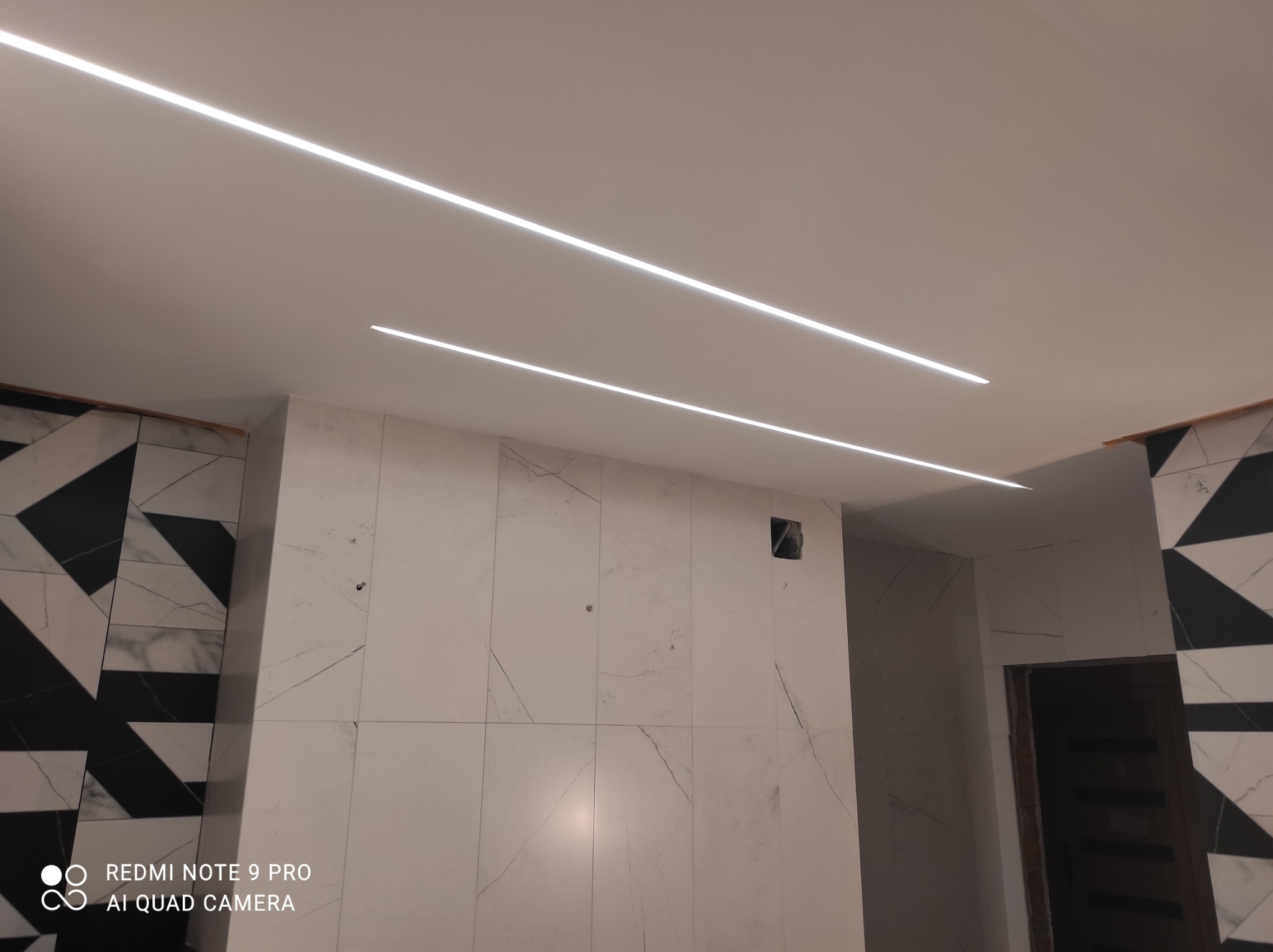 Wykończona łazienka z białymi płytkami imitującymi marmur, czarno-białą mozaiką oraz nowoczesnym oświetleniem LED w suficie.