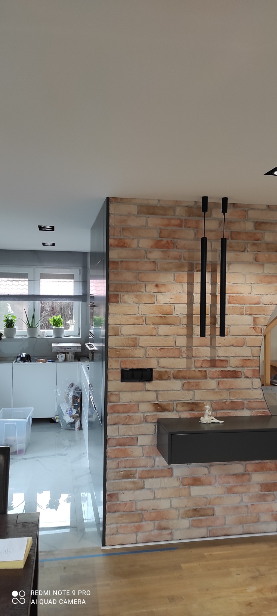 Ceglana ściana dekoracyjna z czarnymi lampami wiszącymi i szarą szafką w nowoczesnym wnętrzu, widok na kuchnię z białymi szafkami i roślinami w doniczkach na parapecie.
