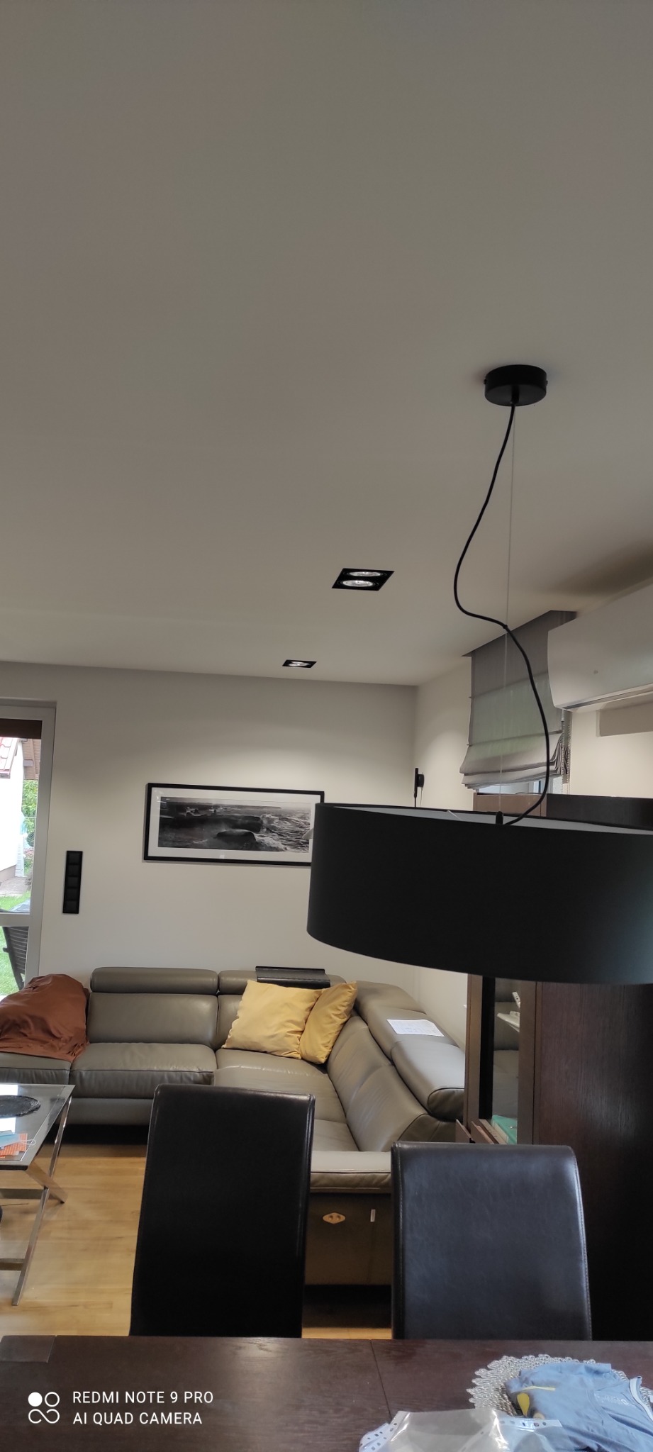 Nowoczesny salon z szarą skórzaną sofą narożną, żółtymi poduszkami, czarnym obrazem w ramie i minimalistycznym oświetleniem sufitowym.