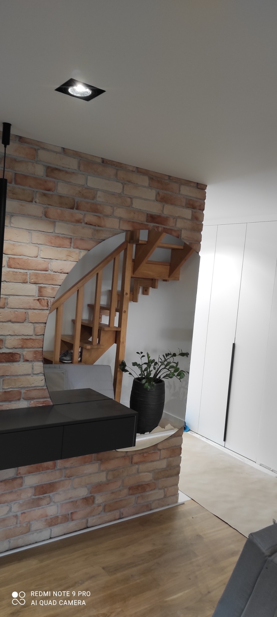 Owalne lustro na ceglanej ścianie odbija drewniane schody i zieloną roślinę w czarnej donicy, obok biała szafa wnękowa z czarnymi uchwytami.