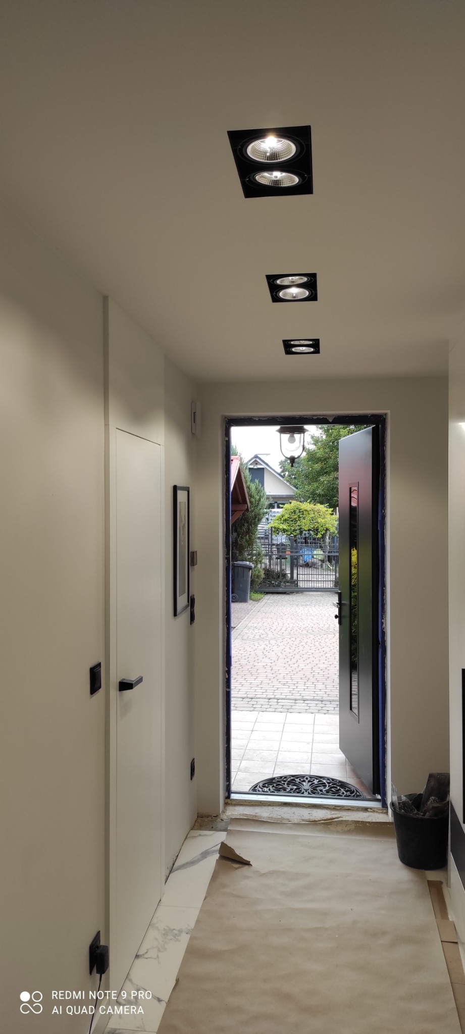 Korytarz w trakcie prac wykończeniowych: położona marmurowa podłoga, zabezpieczona kartonem, widoczne czarne oprawy oświetleniowe sufitowe i otwarte drzwi wejściowe z widokiem na posesję.
