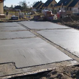 Świeżo wylane, podzielone na sekcje betonowe fundamenty domu jednorodzinnego z wystającymi pionowo rurami kanalizacyjnymi, na tle zabudowy mieszkalnej.