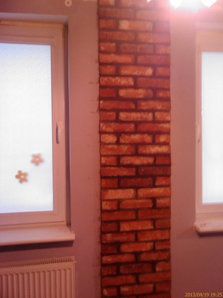 Wąski fragment ściany wykończony dekoracyjną płytką ceglaną w odcieniach czerwieni i brązu, sąsiadujący z oknem z naklejonymi kwiatkami i grzejnikiem, w pomieszczeniu o szarych ścianach.