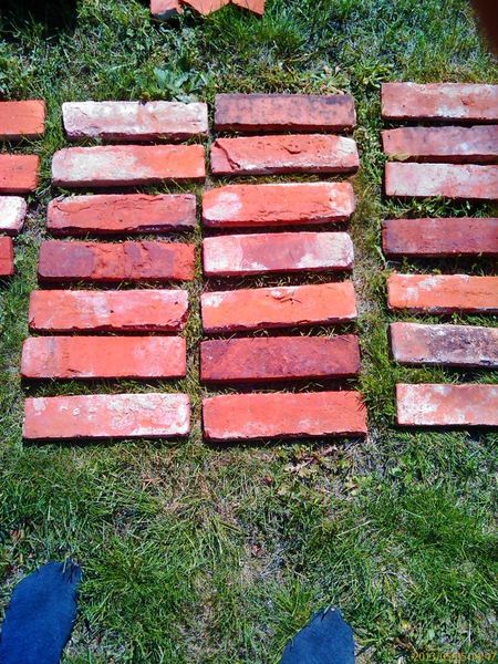 Ułożone na trawie płytki ceglane o zróżnicowanej kolorystyce, od jasnoczerwonej do ciemnobrązowej, z widocznymi fragmentami niebieskich butów w dolnej części kadru.