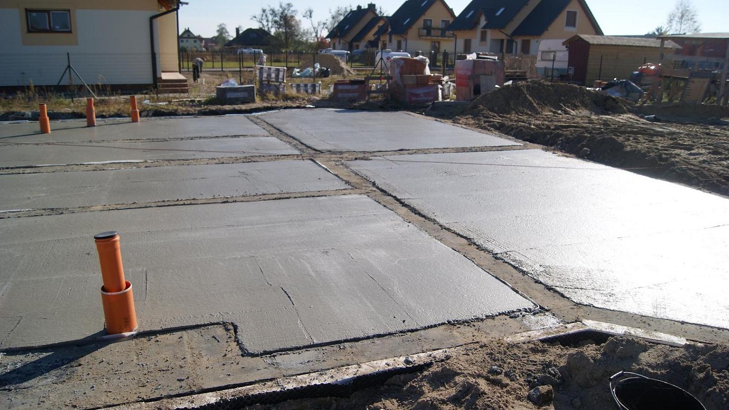 Świeżo wylane, podzielone na sekcje betonowe fundamenty domu jednorodzinnego z wystającymi pionowo rurami kanalizacyjnymi, na tle zabudowy mieszkalnej.