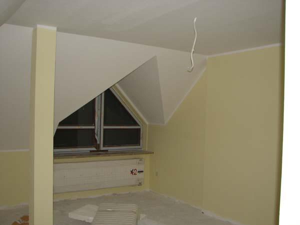 Poddasze w trakcie remontu: ściany pomalowane na jasnożółto, okno trójkątne w szczycie, grzejnik pod oknem, widoczne kable zwisające z sufitu i płyty izolacyjne na podłodze.