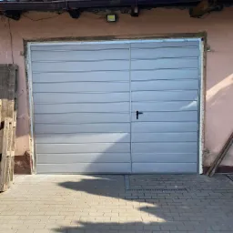 Szara, segmentowa brama garażowa z czarną klamką, zamontowana w różowej elewacji budynku, widok z zewnątrz na brukowany podjazd.