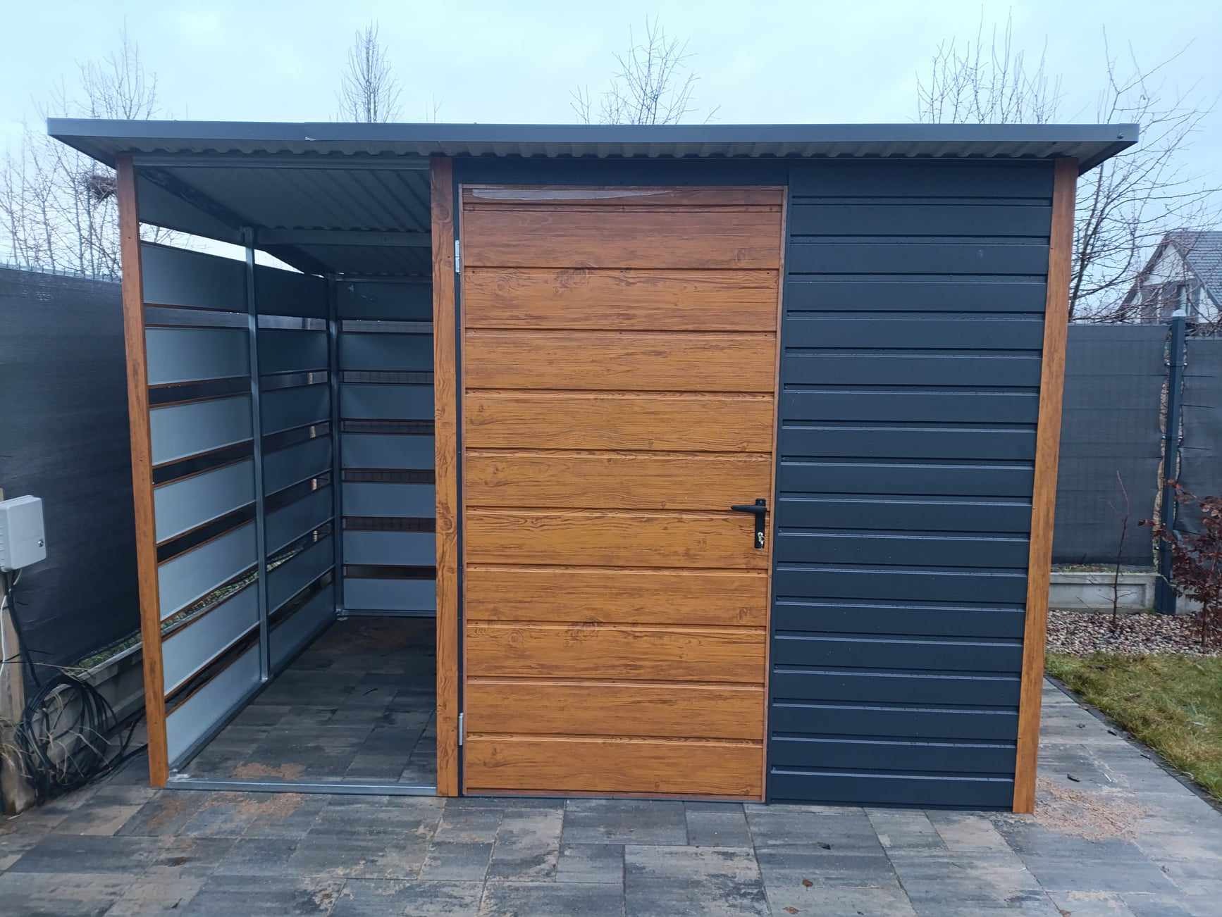 Metalowy garaż blaszany z drewnopodobnymi drzwiami, widoczne elementy konstrukcyjne i posadzka z kostki brukowej na zewnątrz.