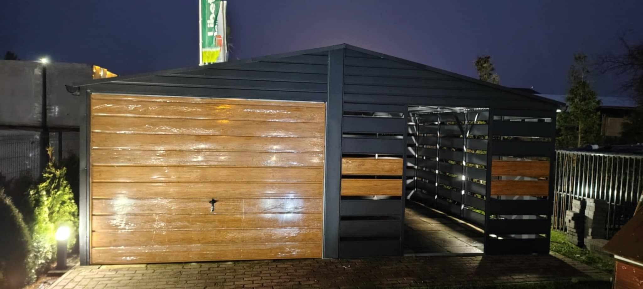 Nowoczesny garaż blaszany z bramą segmentową o drewnianym wykończeniu i ażurową ścianą boczną, oświetlony lampą zewnętrzną na posesji z kostki brukowej, sfotografowany nocą.