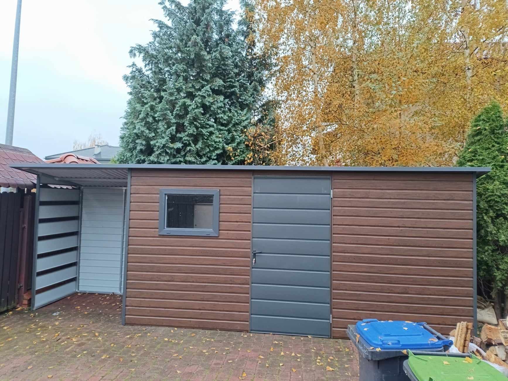 Nowoczesny brązowy garaż blaszany z szarą bramą i oknem, widoczne kosze na śmieci i jesienne drzewa w tle.