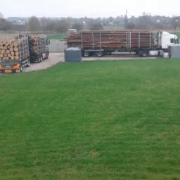 Dwa samochody ciężarowe załadowane okrąglakami, jeden z dźwigiem HDS, stoją na placu obok trawnika.