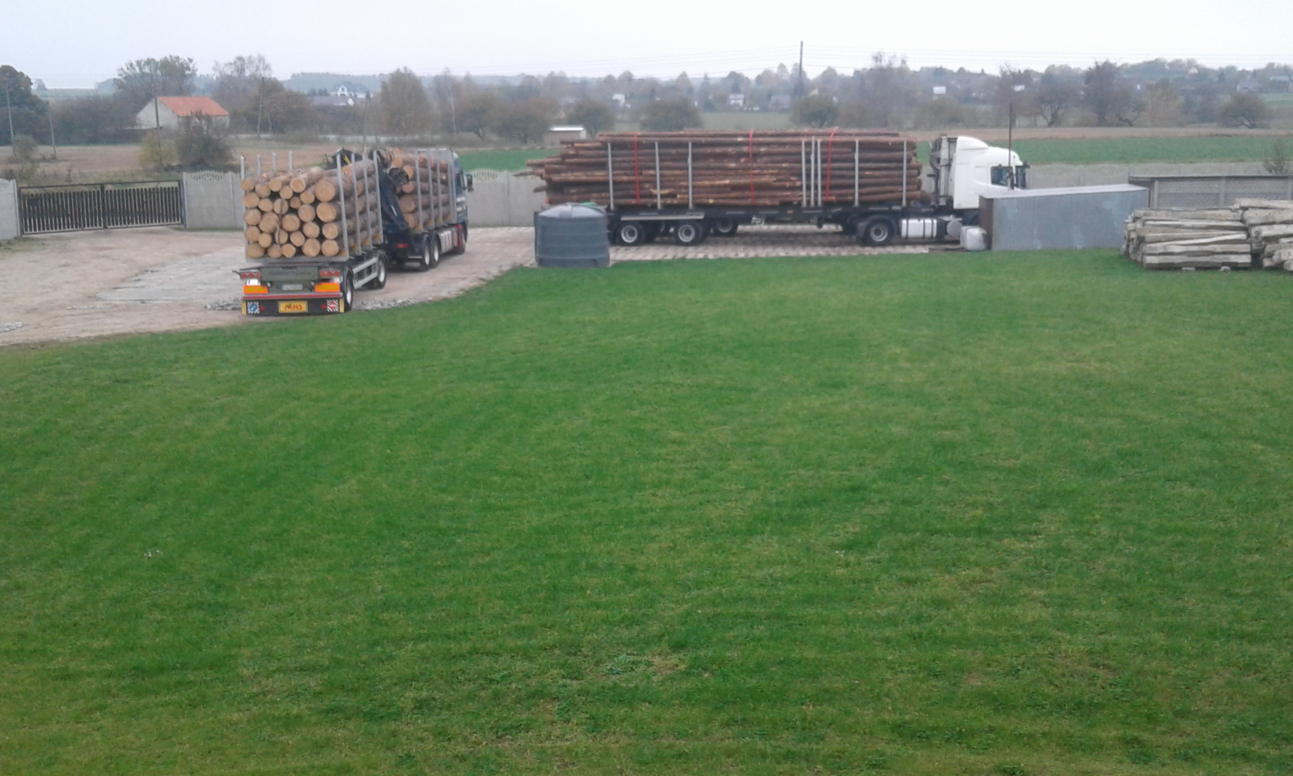 Dwa samochody ciężarowe załadowane okrąglakami, jeden z dźwigiem HDS, stoją na placu obok trawnika.