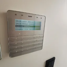 Panel sterowania alarmem Satel z podświetlonym ekranem dotykowym, wskazującym na zamknięte rolety w salonie i kuchni, zamontowany na jasnej ścianie obok czarnego włącznika.