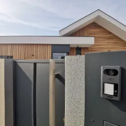 Szary wideodomofon BCS zamontowany na metalowej bramie wjazdowej, z nowoczesnym domem z drewnianą elewacją w tle.