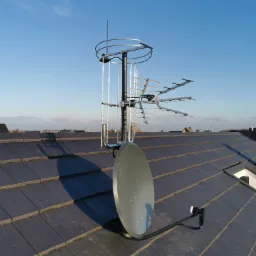 Antena satelitarna i antena telewizyjna zamontowane na dachu domu pokrytym ciemną dachówką, widok z góry na tle błękitnego nieba.