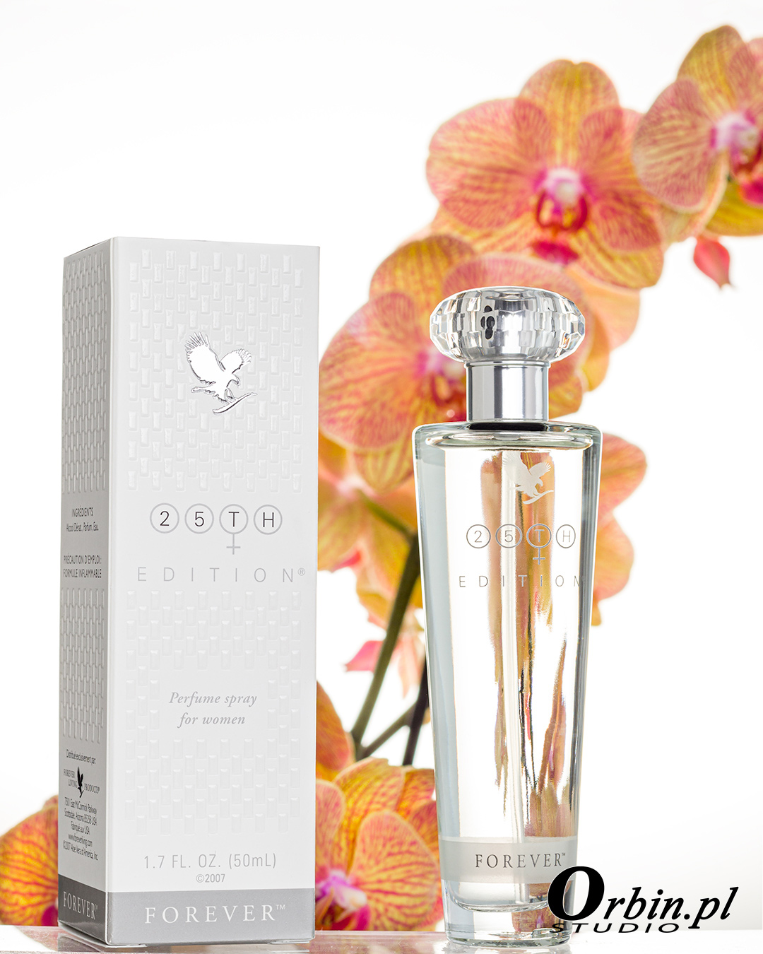 Flakon perfum '25th Edition' marki Forever, ustawiony obok pudełka na tle rozmytych, pomarańczowych orchidei. Flakon ma przezroczysty korpus i kryształową nakrętkę.