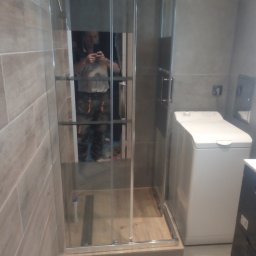 Stel-BUD - Nowoczesna kabina prysznicowa z przesuwnymi drzwiami, widoczna pralka i odbicie fotografa w szkle, wykończenie łazienki w szarościach z drewnopodobną podłogą.