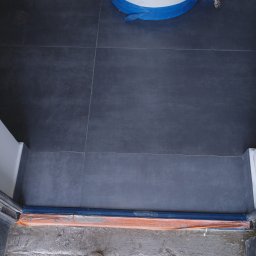 Stel-BUD - Nowa, ciemnoszara podłoga z płytek w łazience, z widocznym bojlerem zabezpieczonym folią oraz fragmentem buta na nierównej posadzce przed wejściem.