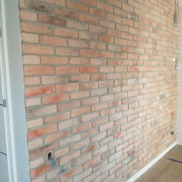 Stel-BUD - Ściana z cegieł dekoracyjnych w odcieniach czerwieni i beżu, z widocznym otworem na gniazdko elektryczne i fragmentem białej listwy przypodłogowej oraz ościeżnicy drzwiowej.