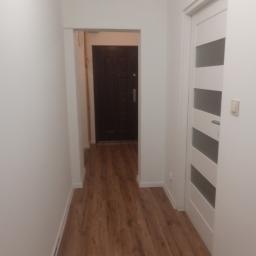 Stel-BUD - Korytarz z panelami podłogowymi imitującymi drewno, białe ściany i drzwi z przeszkleniami w paski, widok na ciemne drzwi wejściowe.