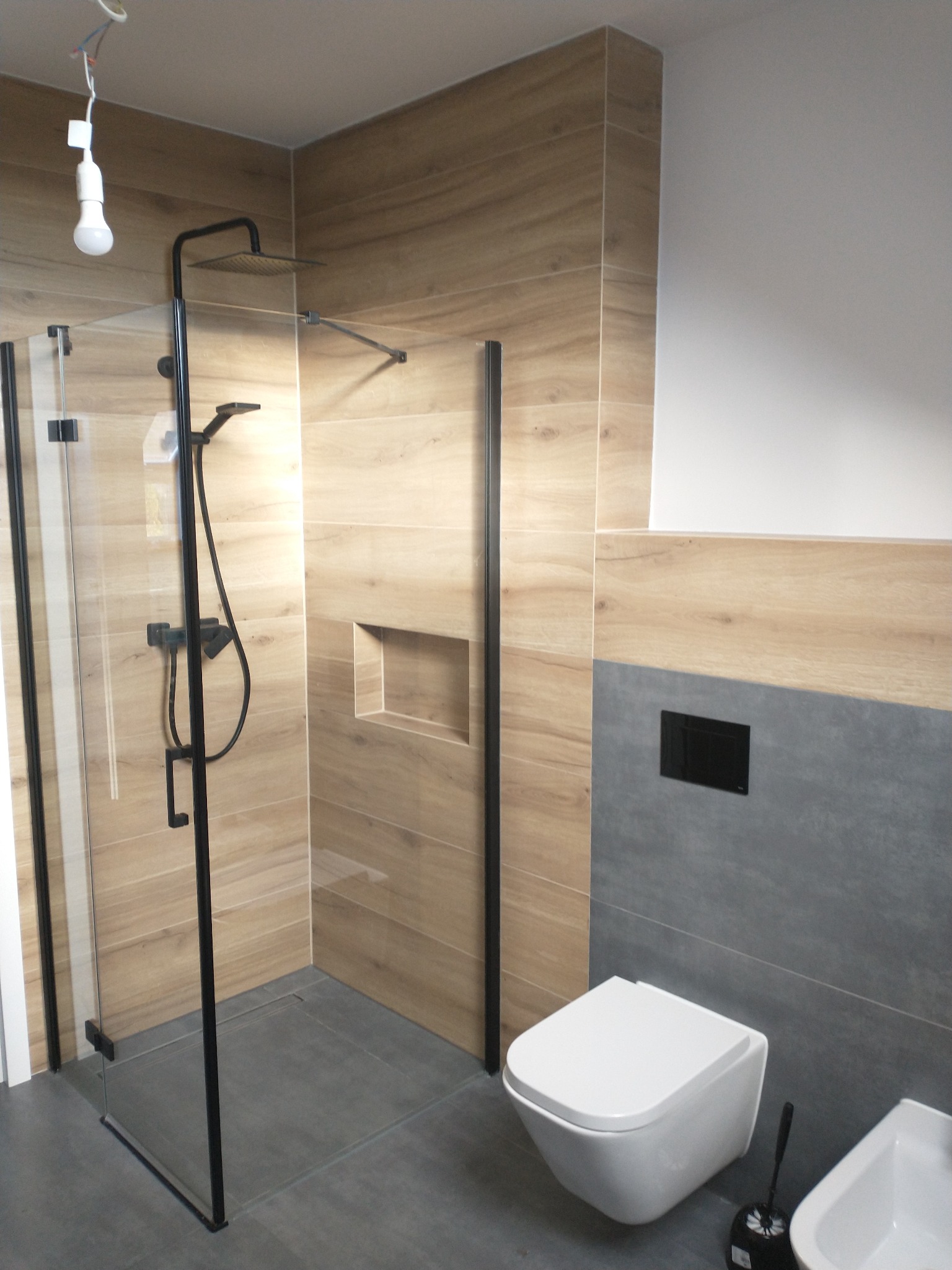 Nowoczesna łazienka z kabiną prysznicową typu walk-in z czarnymi profilami, toaletą podwieszaną i płytkami imitującymi drewno oraz szarym gresem.