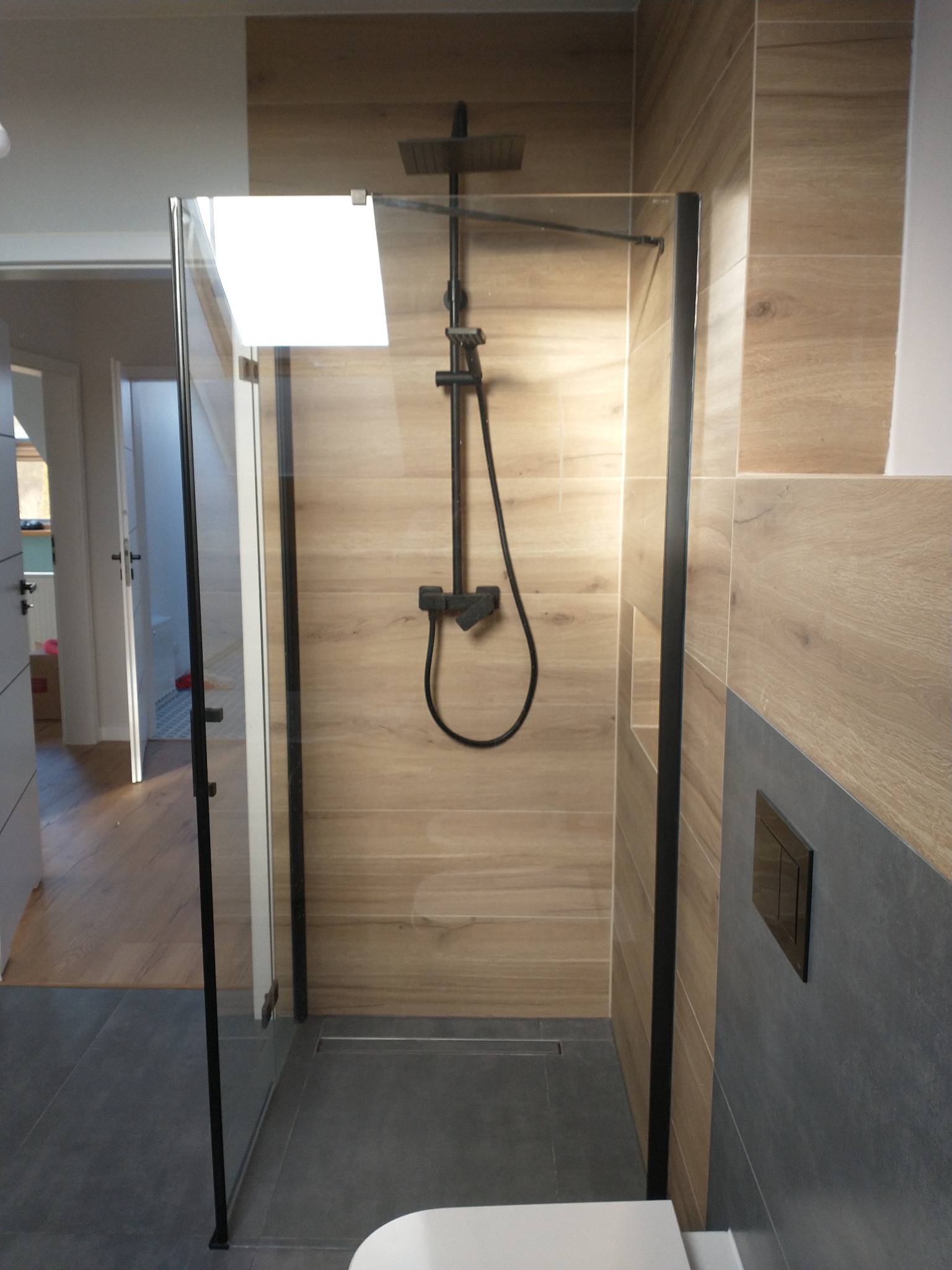 Nowoczesna kabina prysznicowa typu walk-in z czarnymi profilami, drewnopodobne płytki na ścianach, szara podłoga i częściowo szare płytki, widoczny fragment toalety, minimalistyczny design.