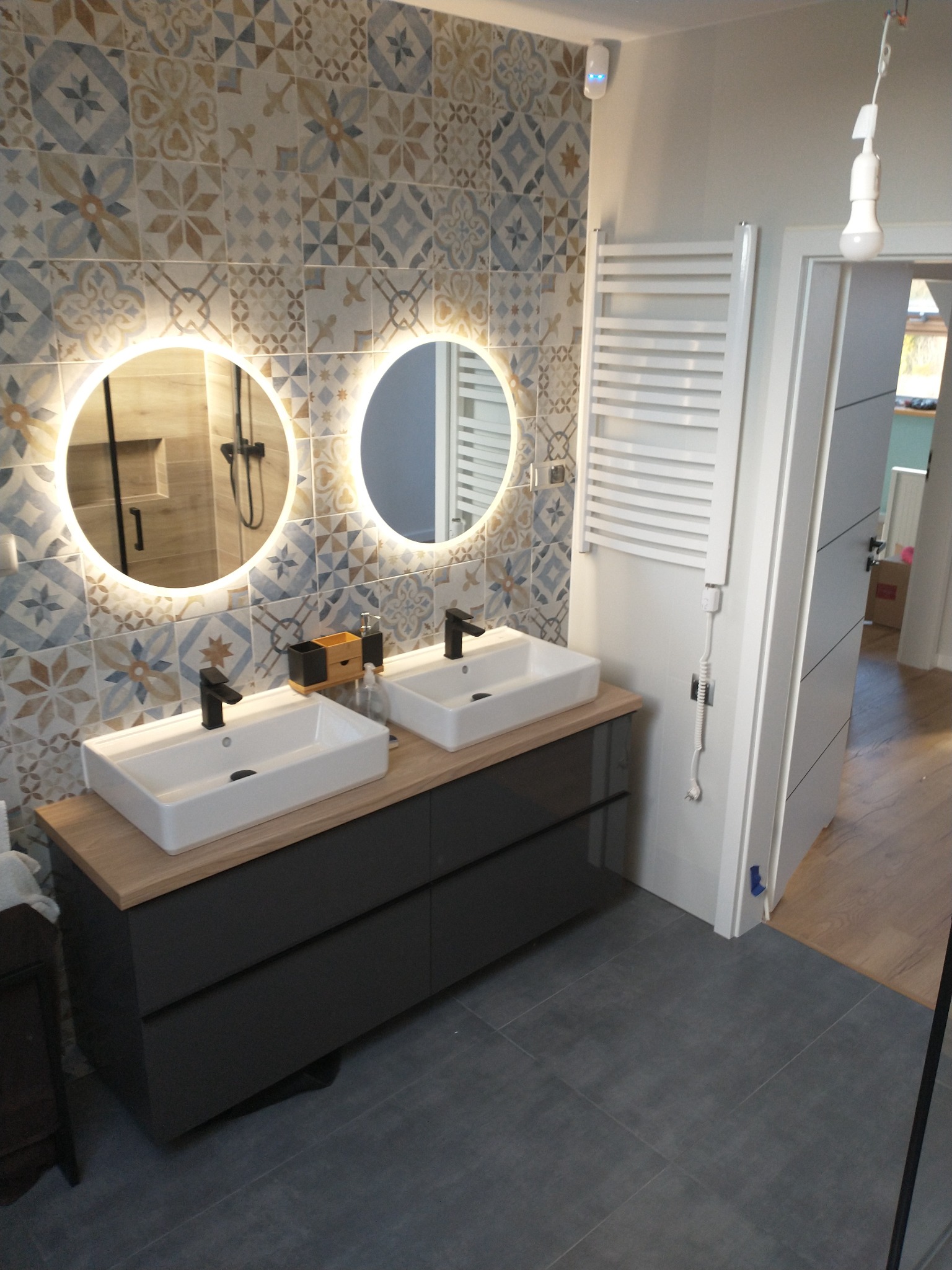 Nowoczesna łazienka z dwoma umywalkami nablatowymi, podświetlanymi okrągłymi lustrami i dekoracyjnymi płytkami ściennymi w odcieniach beżu i błękitu. Widoczny grzejnik drabinkowy i szara podłoga.