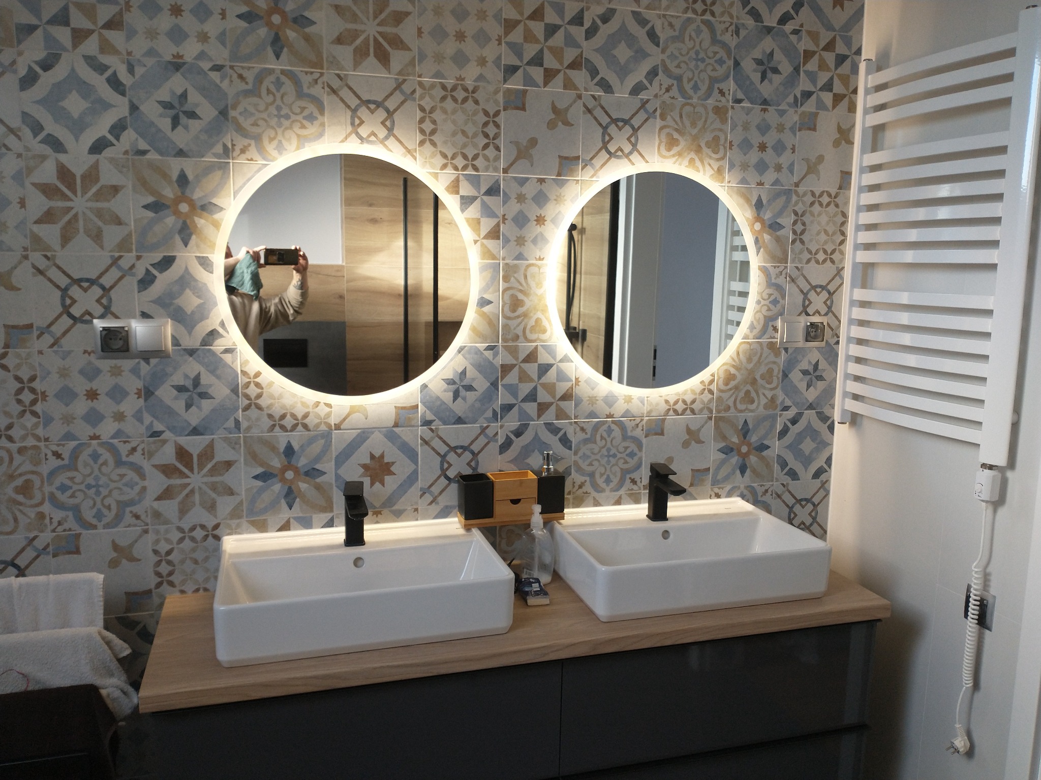 Nowoczesna łazienka z dwoma umywalkami nablatowymi, okrągłymi lustrami z podświetleniem LED i dekoracyjnymi płytkami ściennymi w stylu patchwork. Widoczny grzejnik drabinkowy i armatura w kolorze...