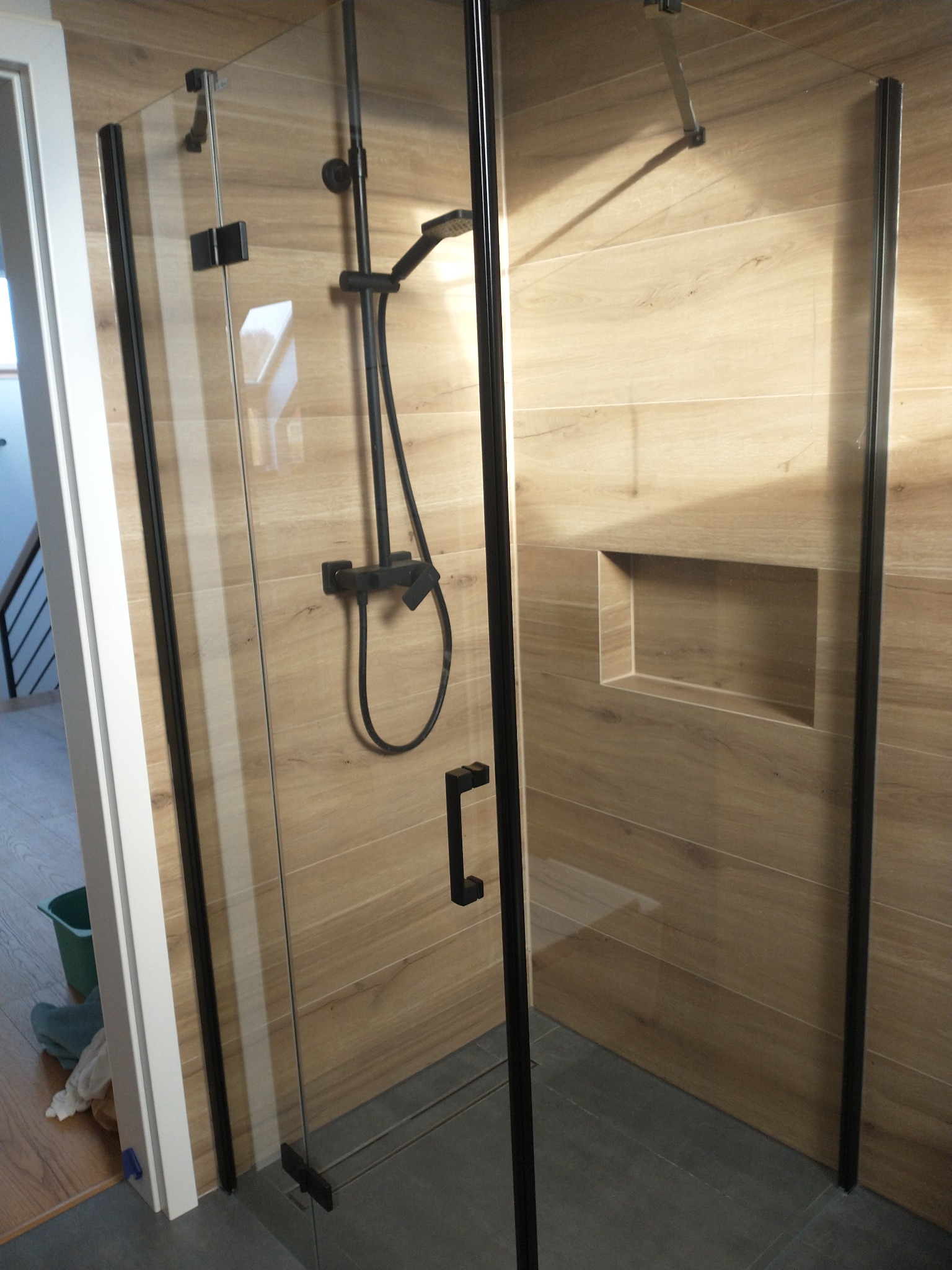 Nowoczesna kabina prysznicowa z czarnymi profilami i deszczownicą na tle ściany z płytek imitujących drewno, z wnęką na kosmetyki.