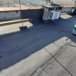 Płaski dach pokryty nową, ciemną papą termozgrzewalną, widoczny komin, klimatyzator oraz butla z gazem i szczotka dekarska.