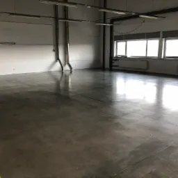 Błyszcząca, szara posadzka w hali przemysłowej z widoczną żółtą linią wyznaczającą strefę, oświetlona przez okna i lampy sufitowe.