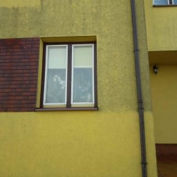 alpinmax.pl - Fragment żółtej elewacji budynku z brązową cegłą, okno z białymi roletami i widocznymi zabrudzeniami na szybie, rynna odprowadzająca wodę.