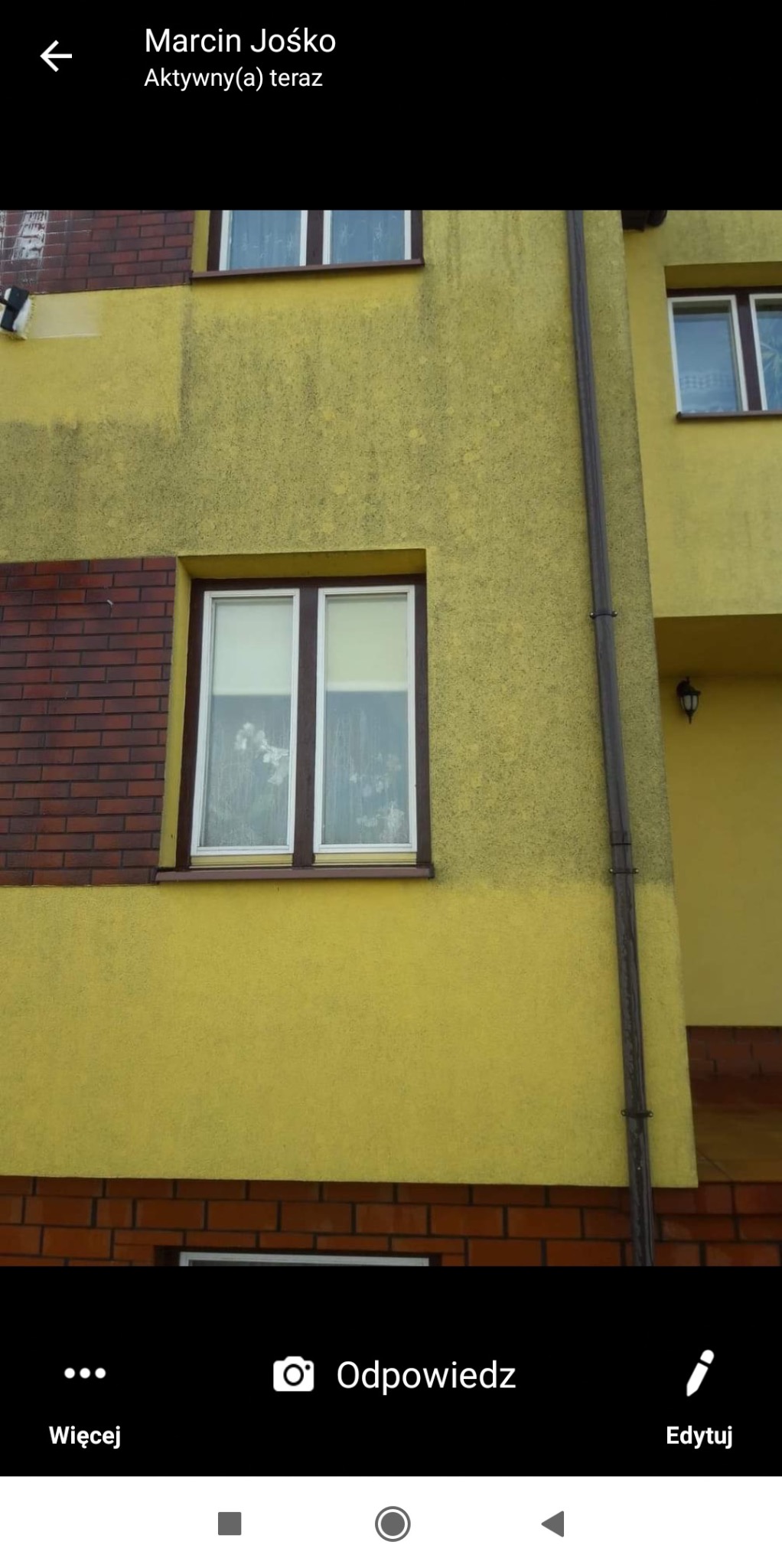 Fragment żółtej elewacji budynku z brązową cegłą, okno z białymi roletami i widocznymi zabrudzeniami na szybie, rynna odprowadzająca wodę.