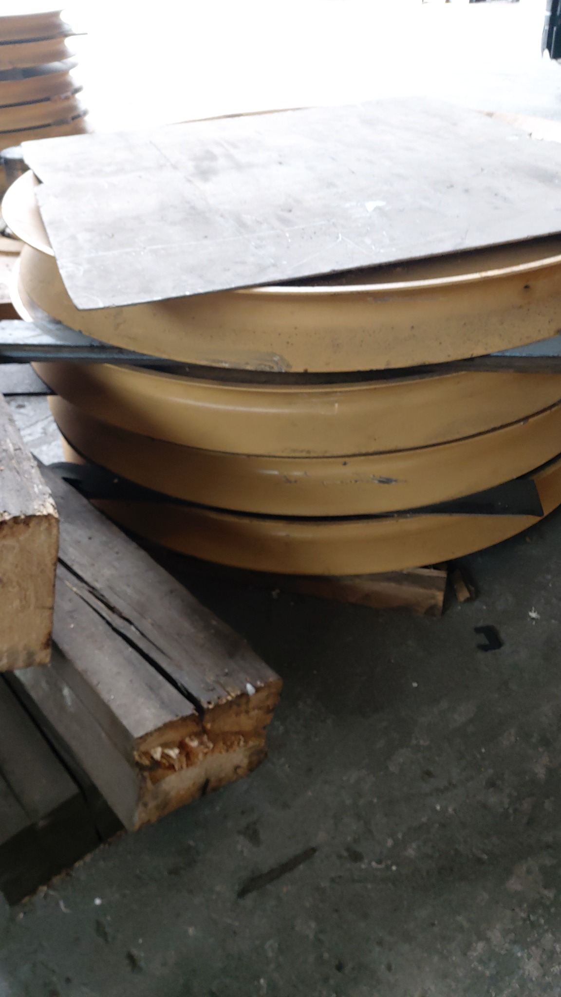 Stos żółtych, okrągłych elementów z metalu, prawdopodobnie obręczy, ułożonych na drewnianych belkach, z metalową płytą na wierzchu, w hali produkcyjnej.