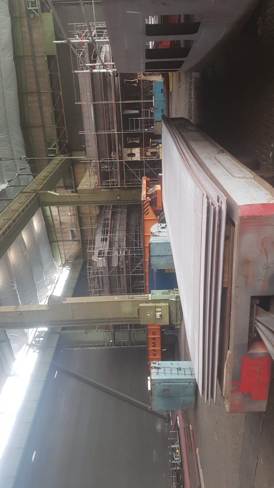 Wnętrze hali produkcyjnej z ułożonymi płaskimi elementami metalowymi, widoczne rusztowania i suwnica w tle.