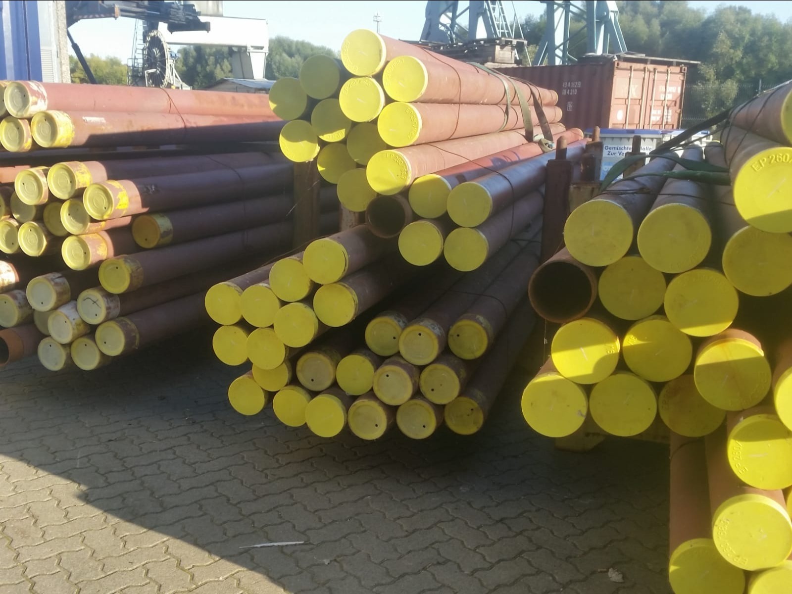 Stosy rur stalowych z żółtymi zaślepkami na końcach, składowane na placu budowy; widoczny kontener i dźwig w tle.
