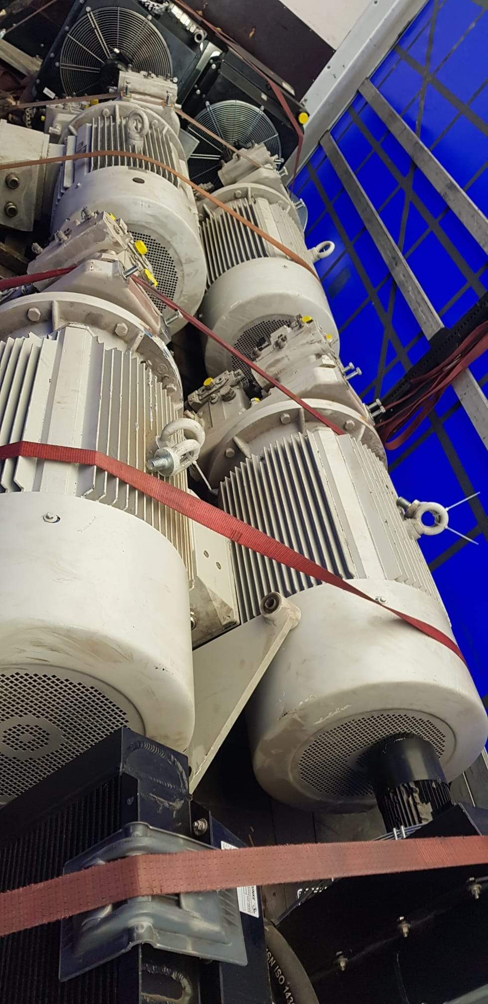 Trzy duże, szare silniki elektryczne z żeberkową obudową, zabezpieczone czerwonymi pasami transportowymi, umieszczone na konstrukcji z niebieskim tłem.