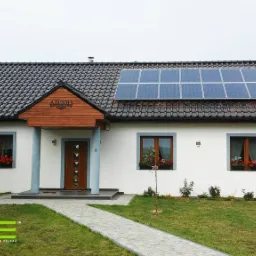 Dom jednorodzinny z panelami słonecznymi na dachu, widoczne okna z czerwonymi kwiatami w skrzynkach, numer domu 'Złota 52' oraz rok budowy 'A.D. 2013' nad wejściem.