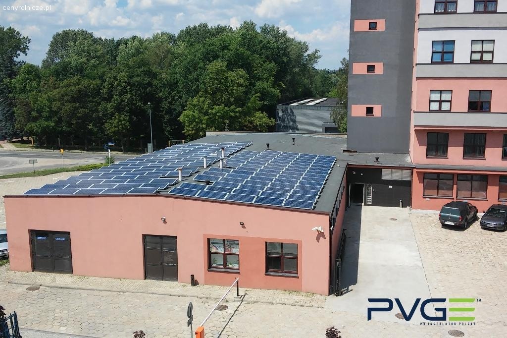 Widok z góry na budynek z zainstalowanymi panelami słonecznymi na dachu, obok budynek mieszkalny, zaparkowane samochody i zieleń w tle.