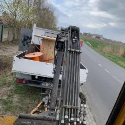 Widok z kabiny koparki na wywrotkę z drewnianymi bębnami na kable i inne odpady, stojącą obok drogi na tle p&oacute;l i zabudowań wiejskich.