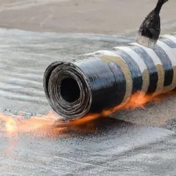 Rolka papy termozgrzewalnej z palnikiem podczas procesu zgrzewania na dachu, widoczne płomienie i stopiony materiał.