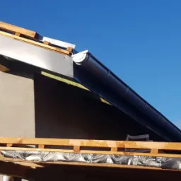Montaż czarnej rynny dachowej na tle błękitnego nieba, widoczne drewniane elementy konstrukcji dachu i fragment ocieplonej ściany budynku.