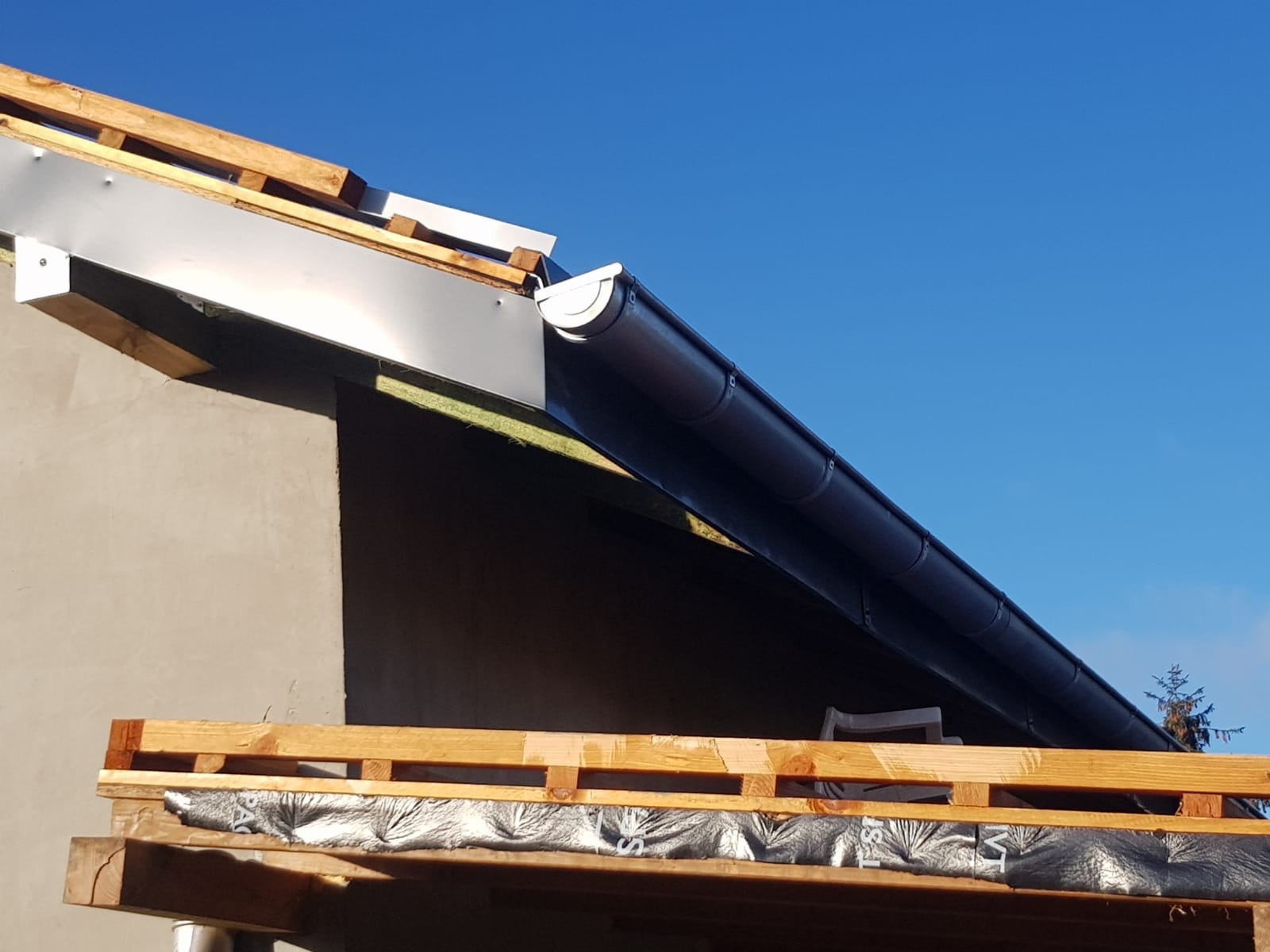 Montaż czarnej rynny dachowej na tle błękitnego nieba, widoczne drewniane elementy konstrukcji dachu i fragment ocieplonej ściany budynku.