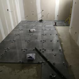 Usługi ogólnobudowlane - Układanie ciemnoszarych płytek na podłodze w surowym pomieszczeniu, widoczne poziomica, paczki z klejem i narzędzia, laser krzyżowy wyznacza linię, oświetlenie budowlane