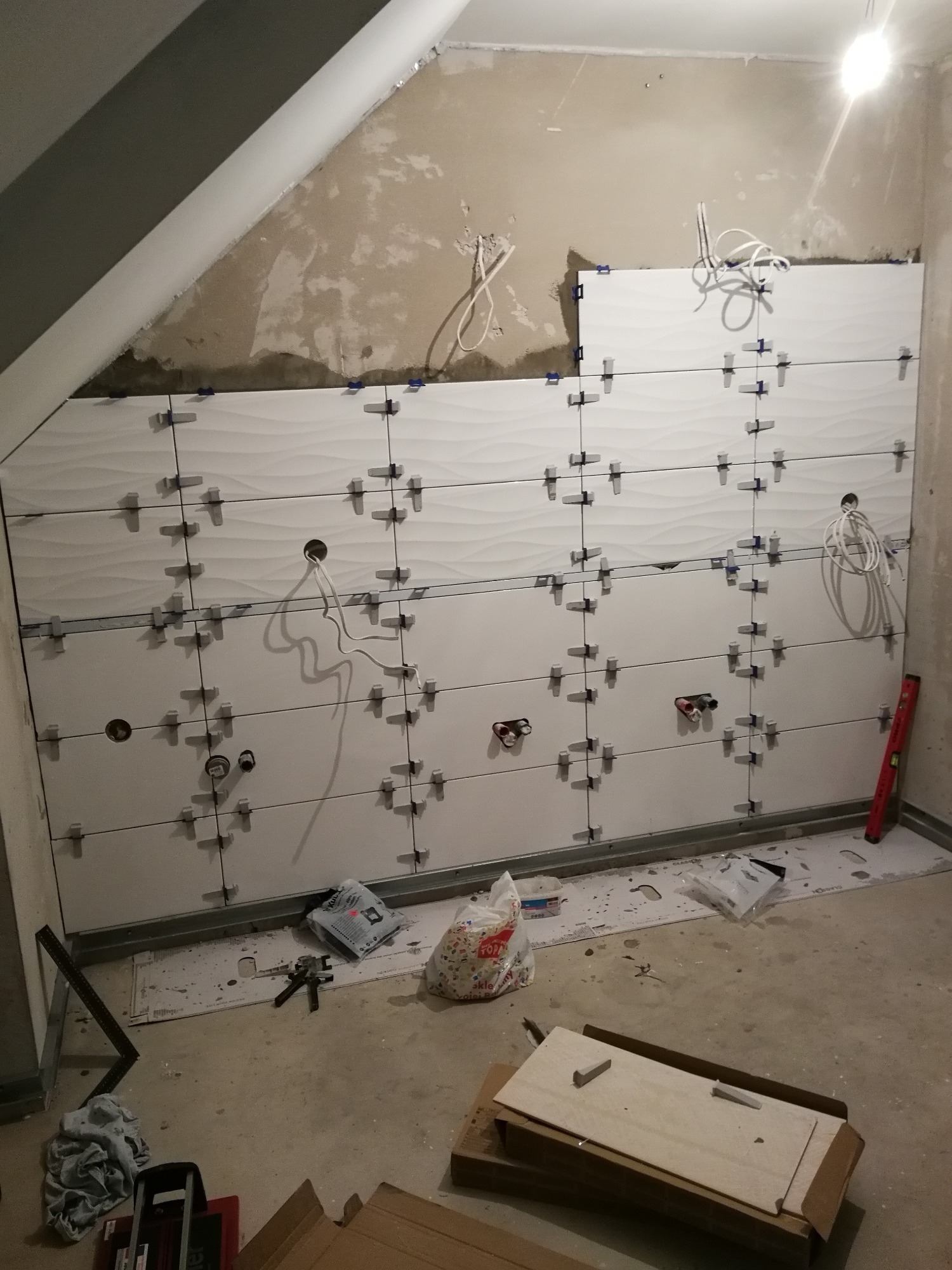 Ściana łazienki w trakcie układania białych, falistych płytek ceramicznych z użyciem krzyżyków dystansowych, widoczne otwory na instalacje, poziomica i narzędzia na podłodze.
